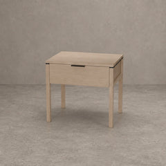 A light natural oak nightstand with a single drawer and a slim black metal handle, set against a textured gray concrete wall and floor.