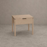 A light oak wood nightstand with a single drawer, featuring a minimalist black metal pull handle and slim black metal corner accents, set against a textured gray concrete wall and floor.