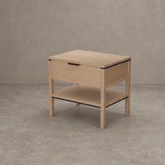 A light oak wood nightstand with a single drawer, open lower shelf, and thin black metal trim accents, set against a textured gray concrete wall and floor.