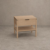 A light oak wood nightstand with a single drawer, an open lower shelf, and a slim black metal handle, set against a textured gray concrete wall and floor.