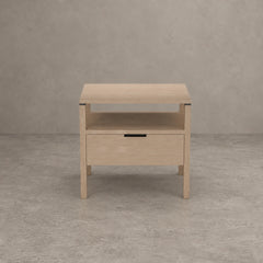 A light natural wood nightstand with a single drawer and an open shelf, set against a textured gray concrete wall and floor.