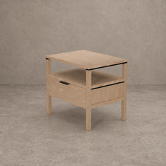 A light oak wood side table with a single drawer, open shelf, and black metal accents, set against a textured gray concrete background.
