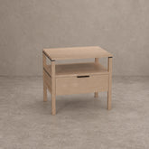 A light natural wood nightstand with a single drawer, open lower shelf, and open top surface, shown against a textured gray concrete-style background.