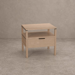 A light natural wood nightstand with a single drawer, open lower shelf, and open top surface, shown against a textured gray concrete-style background.