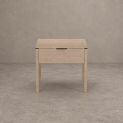 A light natural wood nightstand with a single drawer and a slim black metal pull handle, set against a textured gray concrete wall and floor.