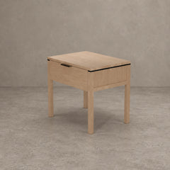 A light oak wood nightstand with a single drawer, featuring a black metal handle and a thin black accent line along the top edge, set against a textured gray concrete wall and floor.