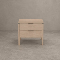 A light oak wood two-drawer nightstand with black metal handles, shown from a front-facing perspective against a textured gray concrete wall and floor.