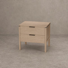 A light oak wood two-drawer nightstand with black metal drawer pulls, shown on a textured gray concrete floor against a matching gray wall.
