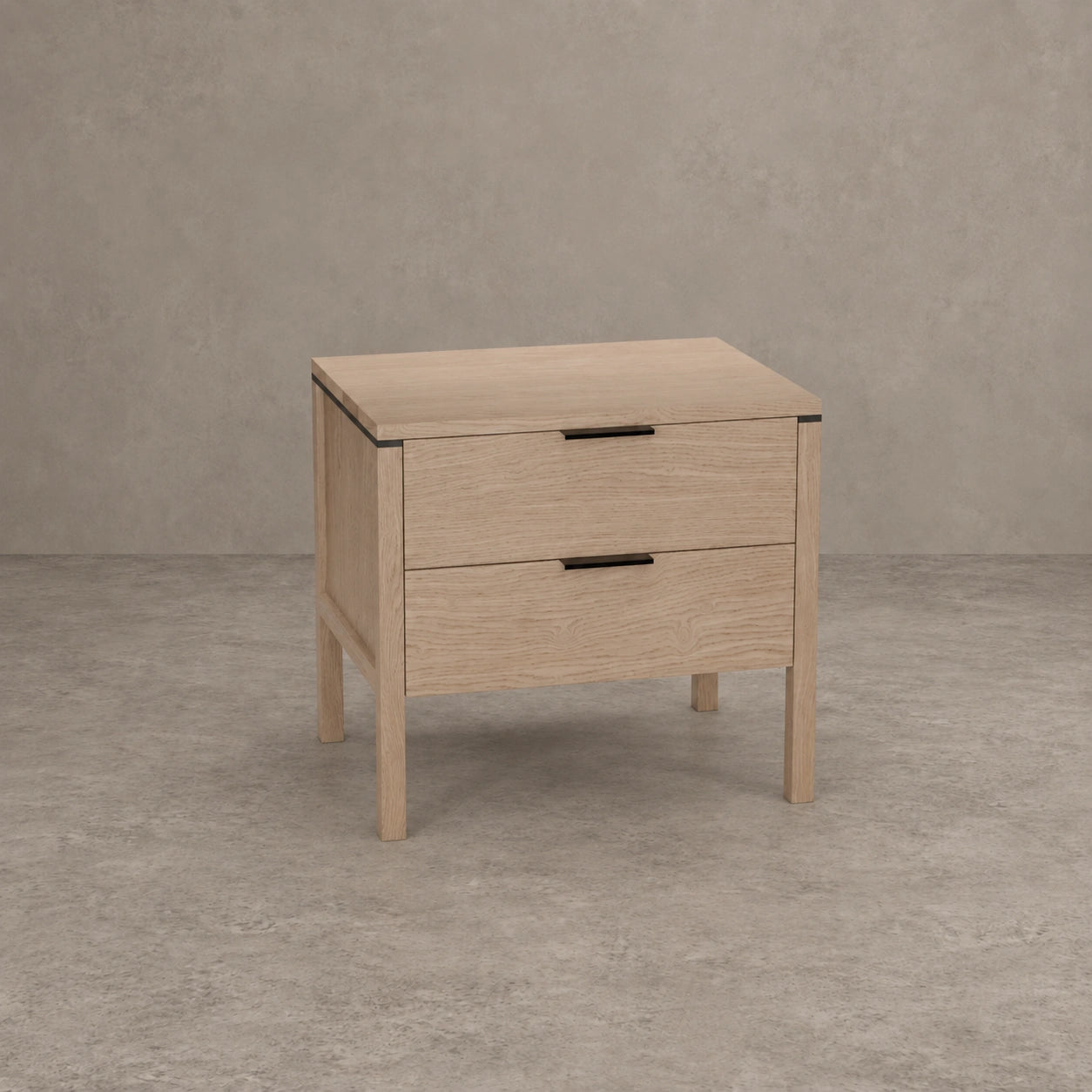 A light oak wood two-drawer nightstand with black metal drawer pulls, shown in a minimalist studio setting with a textured gray concrete floor and wall.