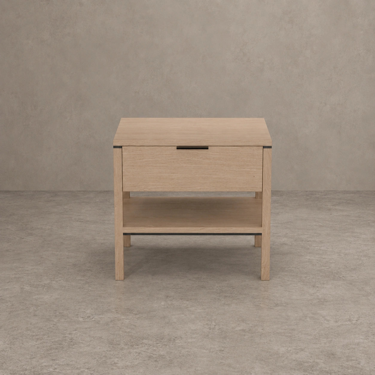 A light oak wood nightstand with a single drawer, an open lower shelf, and a slim black metal handle, set against a textured gray concrete wall and floor.