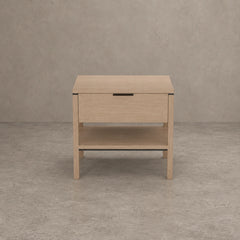 A light oak wood nightstand with a single drawer, an open lower shelf, and a slim black metal handle, set against a textured gray concrete wall and floor.
