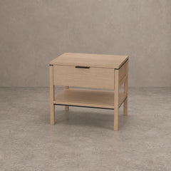 A light oak wood nightstand with a single drawer, open lower shelf, and slim black metal handle, set against a textured gray concrete wall and floor.