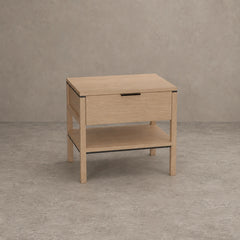 A light oak wood nightstand with a single drawer, an open lower shelf, and a slim black metal handle, set against a textured gray concrete wall and floor.
