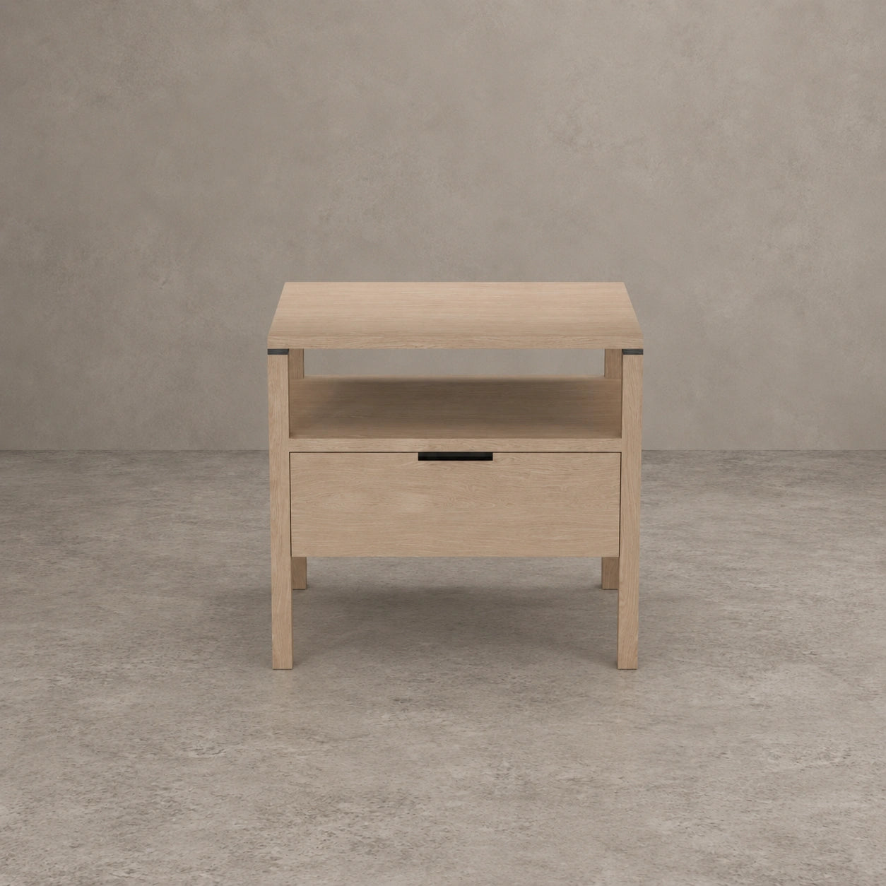 A light natural wood nightstand with a single drawer and an open shelf, set against a textured gray concrete wall and floor.