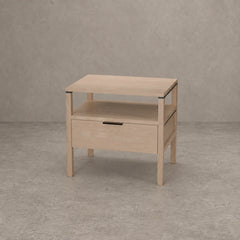 A light natural wood nightstand with a single drawer, open shelf, and open top surface, shown against a textured gray concrete wall and floor.